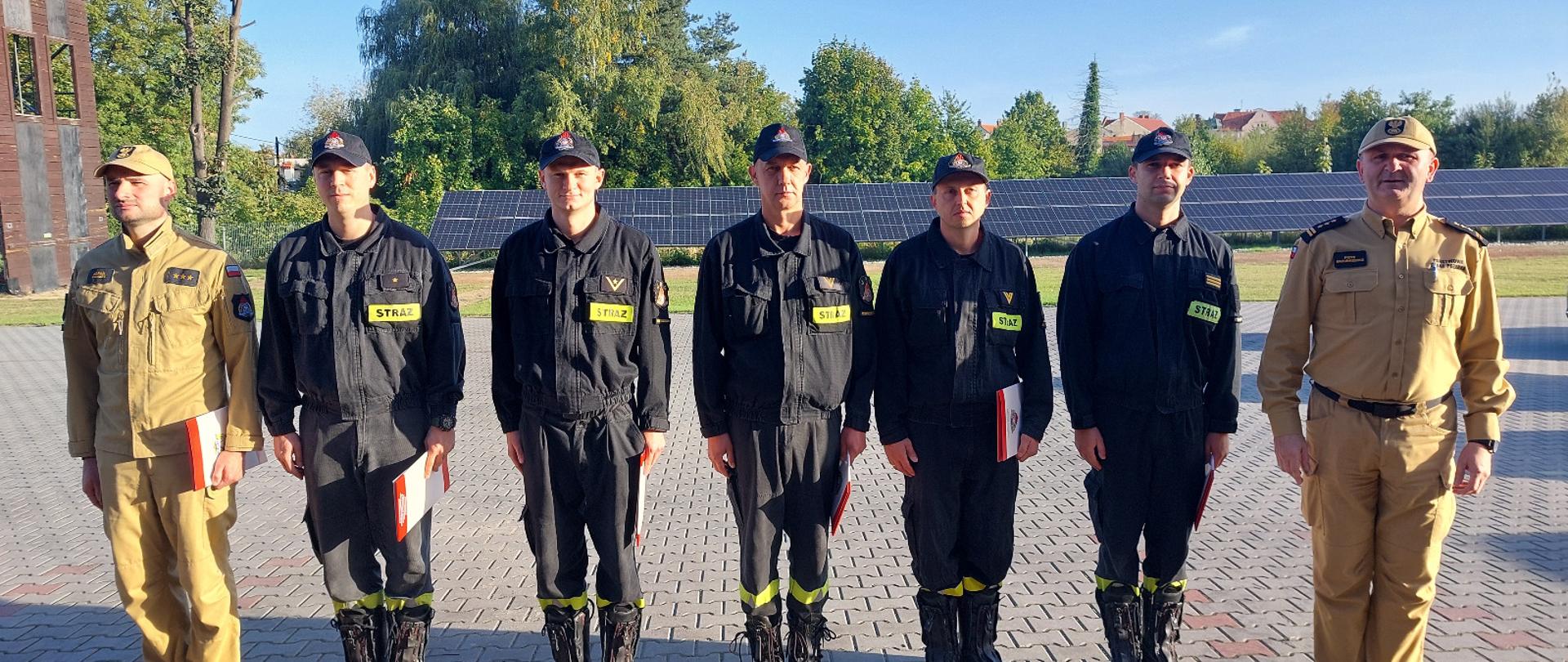 Strażacy stoją w umundurowaniu służbowym na placu wewnętrznym. Na drugim planie widać panele fotowoltaiczne oraz drzewa.