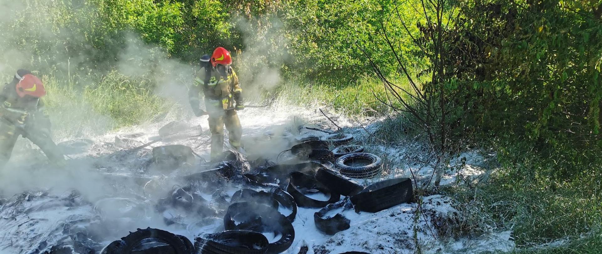 Gorzelnia - Pożar opon w lesie - spalone opony pokryte pianą gaśniczą, widać zadymienie i 2 strażaków z aparatami ochrony dróg oddechowych