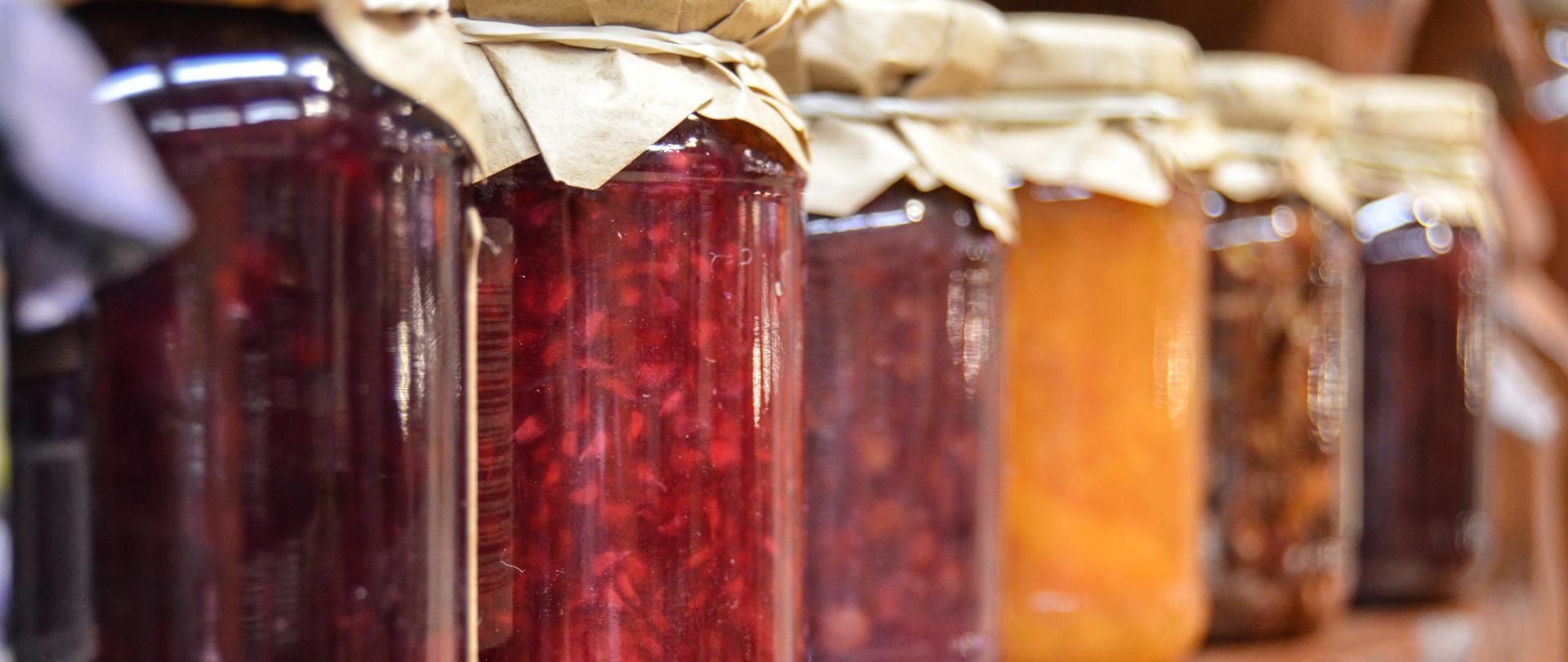 Fotografia przedstawiająca słoiki z przetworami owocowymi ustawione na półce drewnianej w rzędzie