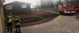 Kolorowa fotografia przedstawia widok utwardzonego placu i wejście do budynku mieszkalnego. Przed wejściem do budynku dwóch umundurowanych druhów strażaków w maseczkach ochronnych i rękawiczkach trzyma roznoszone ulotki informacyjne. Po prawej stronie widoczny czerwony pojazd bojowy straży pożarnej. W tle inne budynki i drzewa.