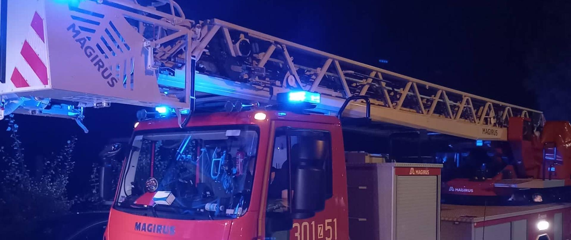 Samochód z drabiną mechaniczną z włączonymi sygnałami świetlnymi, na parkingu przy budynku wielorodzinnym.