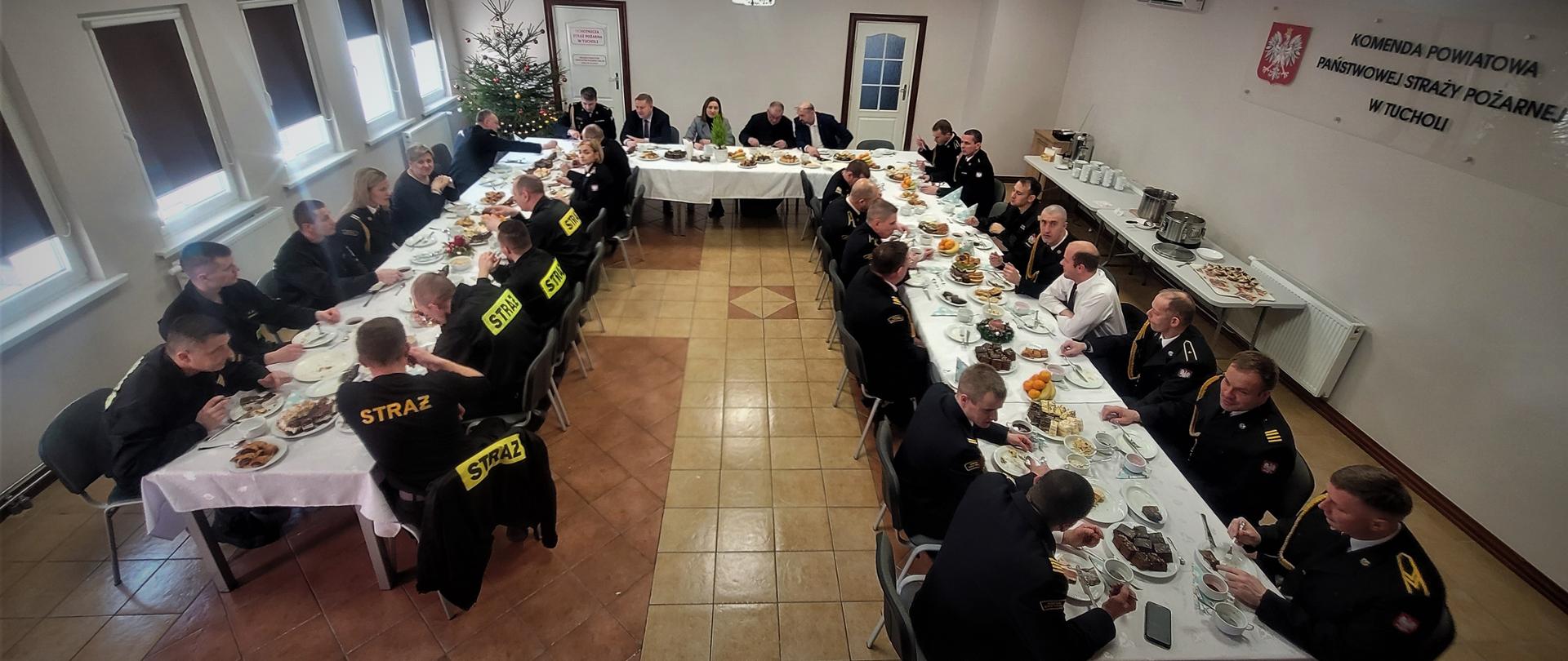 1. Strażacy i cywile siedzą przy stole. Na stołach znajduje się jedzenie. Po lewej stronie jest pięć okien. Po prawej stronie na ścianie widnieje godło Polski oraz napis Komenda Powiatowa Państwowej Straży Pożarnej w Tucholi. W tle widać po prawej i lewej stronie drzwi. Po lewej stronie w rogu stoi choinka. Na środku sufitu wisi rzutnik. Po prawej stronie przy ścianie znajduje się mniejszy stolik z jedzeniem. 