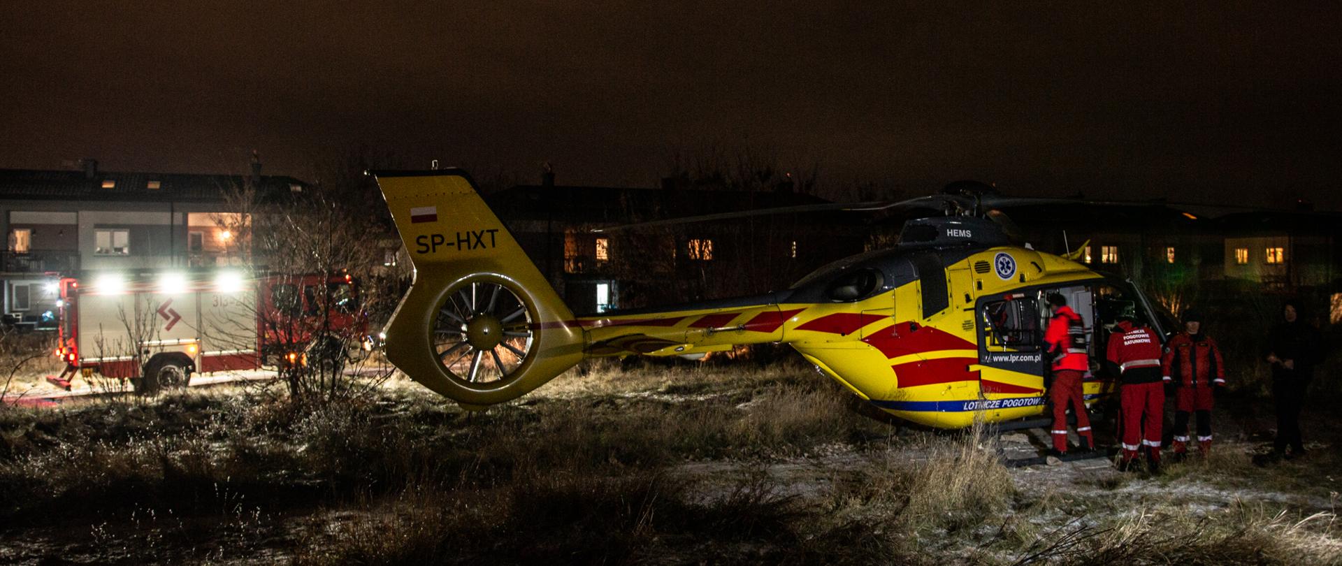 Na zdjęciu na pierwszym planie widoczny jest helikopter LPR i stojących przy nim ratowników medycznych. Na drugim planie w oddali widoczny jest samochód straży pożarnej z włączonym oświetleniem pola pracy.