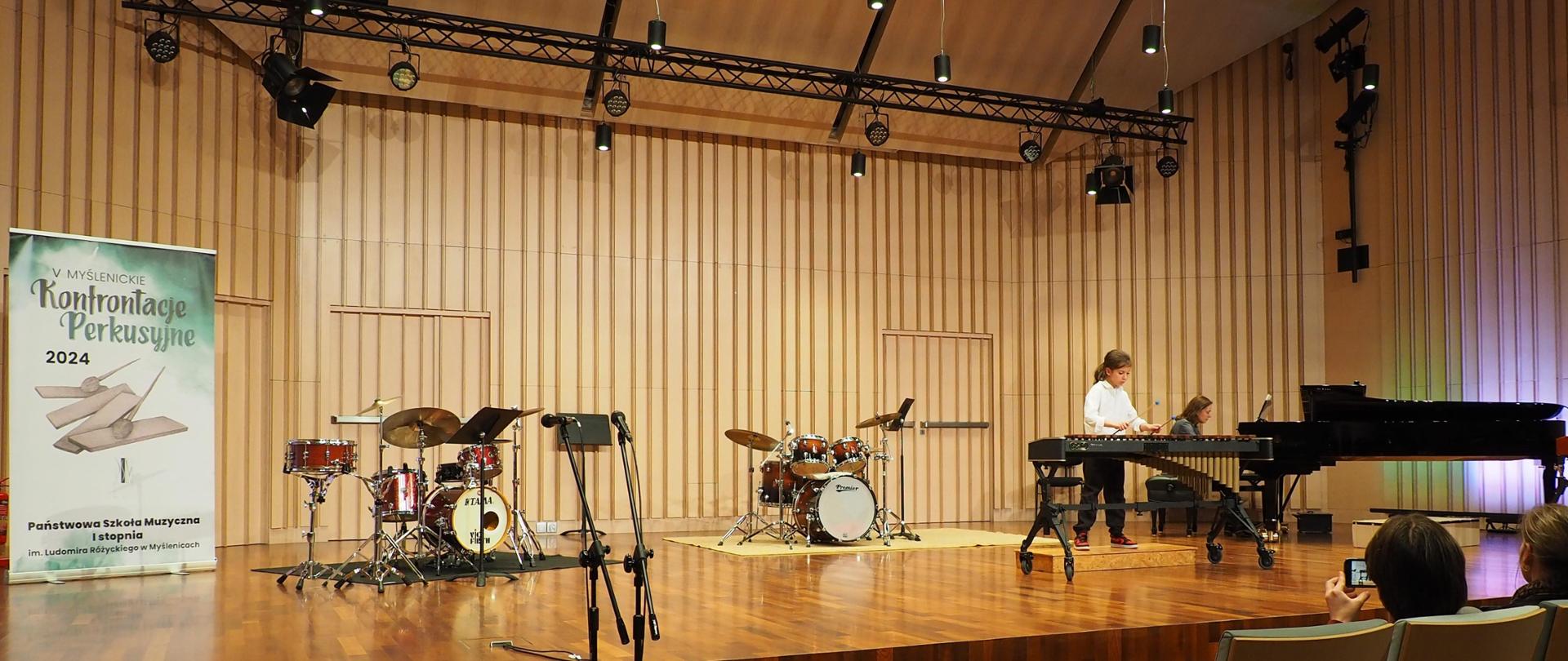 Dwie osoby na scenie w sali koncertowej: uczeń grający na ksylofonie oraz akompaniator, obok dwa zestawy perkusyjne i plakat z napisem: V Myślenickie Konfrontacje Perkusyjne 2024 Państwowa Szkoła I stopnia im. Ludomira Różyckiego w Myślenicach