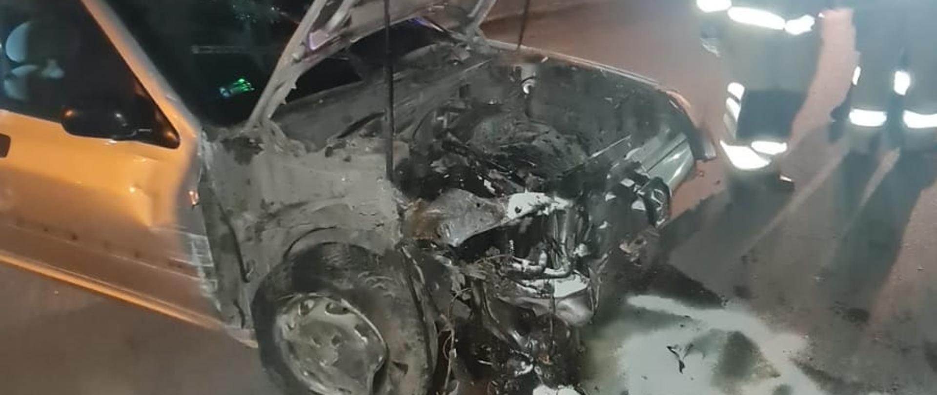 Widać uszkodzony przód samochodu, podniesiona maska, na drodze plama oleju, jest noc