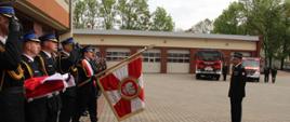 Przegląd pododdziału Zachodniopomorskiego Komendanta Wojewódzkiego PSP, st. bryg. Mirosława Pendera, oddanie honoru pocztów, w tle samochody pożarnicze oraz garaże, niebo całkowicie zachmurzone