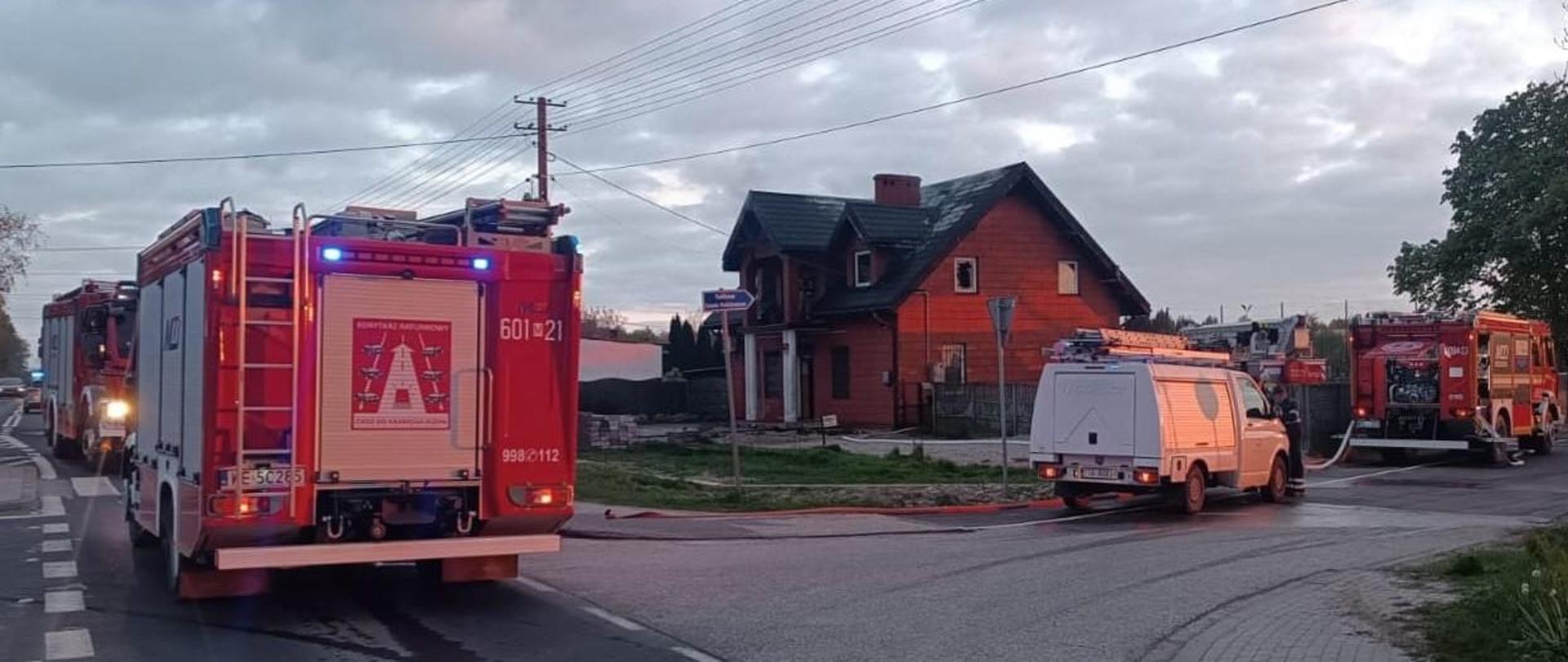 W oddali widać budynek, z którego unosi się dym, co wskazuje na pożar. Na pierwszym planie, na drodze, zaparkowane są czerwone wozy strażackie z włączonymi światłami awaryjnymi, a także biały samochód pogotowia energetycznego. Akcja dzieje się przed zmrokiem.