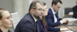 Minister funduszy i polityki regionalnej Grzegorz Puda na posiedzeniu Rady Wykonawczej ds. wdrażania Krajowej Polityki Miejskiej 2030, minister siedzi przy stole i przemawia do mikrofonu, po jego lewej stronie dyrektor Departamentu Strategii MFiPR Renata Calak