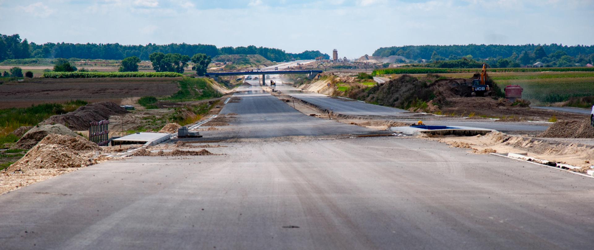 Na zdjęciu widać szeroki, rozległy plac budowy drogi ekspresowej S19 na odcinku między Niedrzwicą Dużą a Kraśnikiem. Ujęcie wykonano z poziomu jezdni, patrząc w dal. Środkowa część kadru to świeżo ułożona, ale jeszcze nieukończona warstwa asfaltu, z wyraźnymi przerwami i fragmentami w trakcie prac. Po obu stronach drogi znajdują się nasypy ziemi, wykopy oraz hałdy piasku i żwiru. W oddali widać maszyny budowlane, m.in. koparkę po prawej stronie, oraz konstrukcję przyszłego wiaduktu. Krajobraz otaczają pola uprawne i pas lasu na horyzoncie. Pogoda jest słoneczna, z lekkim zachmurzeniem. Zdjęcie wykonano w 2021 roku.