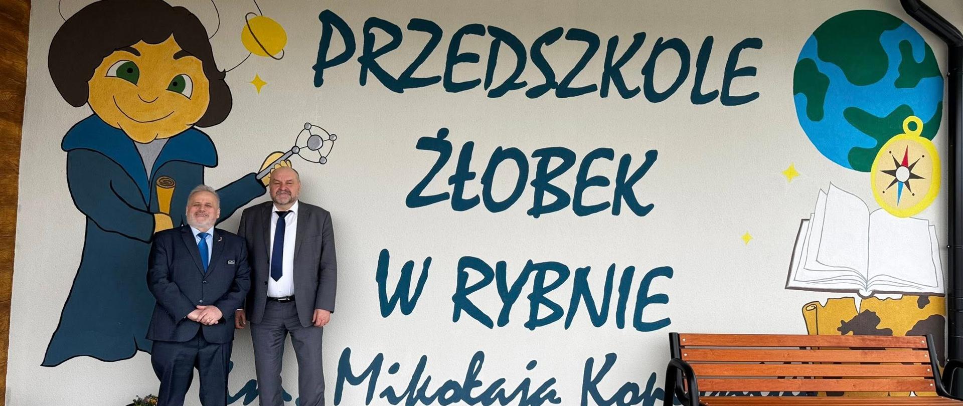 Mężczyźni w garniturach, w tym wicewojewoda, pozują na tle pomalowanej ściany przedszkola