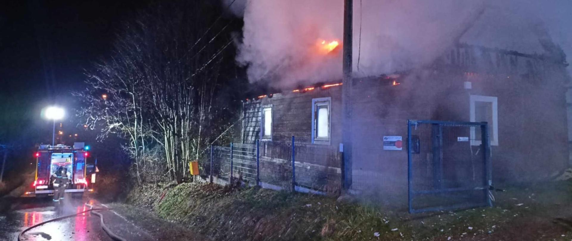 Zdjęcie przedstawia opuszczony budynek mieszkalny, w którym doszło do pożaru. Po lewej stronie, na ulicy rozciągnięte leżą węże gaśnicze, a w głębi stoi samochód strażacki. 