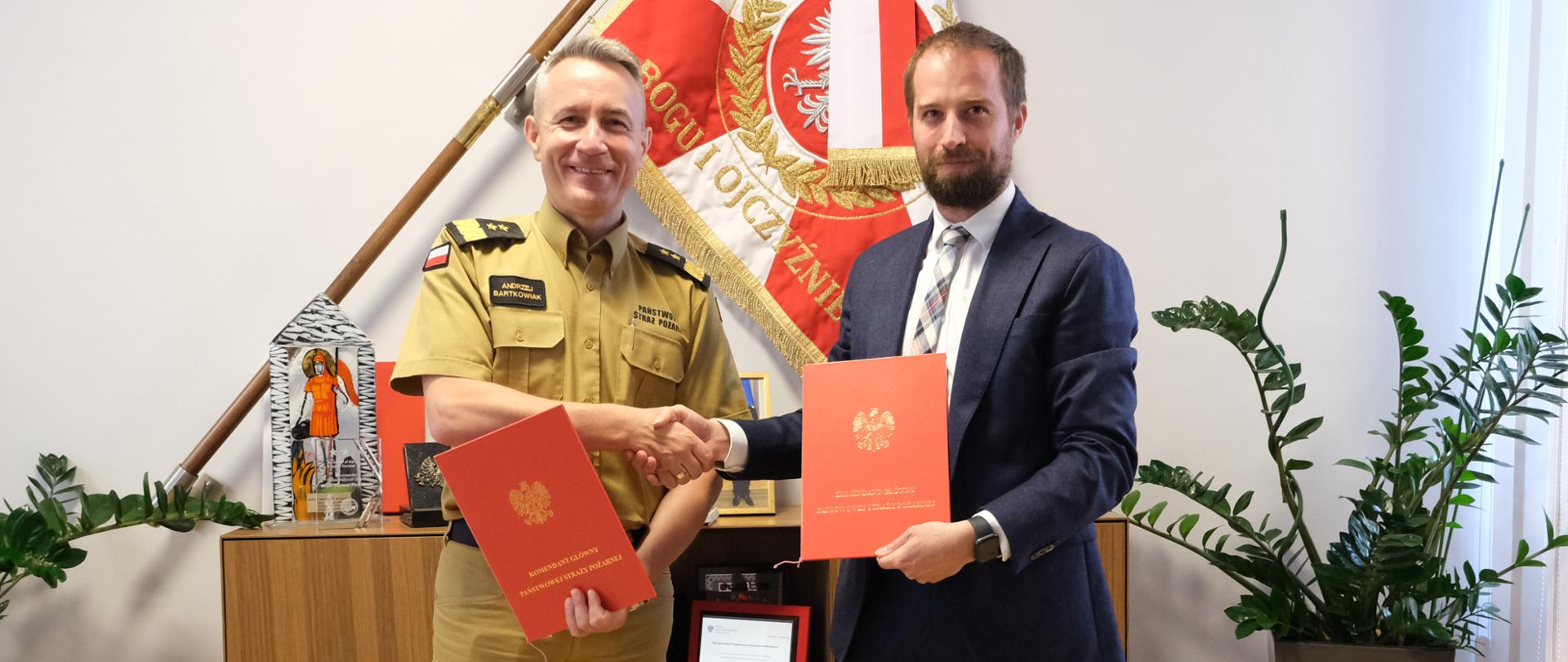 Zdjęcie pamiątkowe po podpisaniu porozumienia. Dwaj mężczyźni trzymający czerwone teczki, na tle sztandaru zawieszonego na ścianie