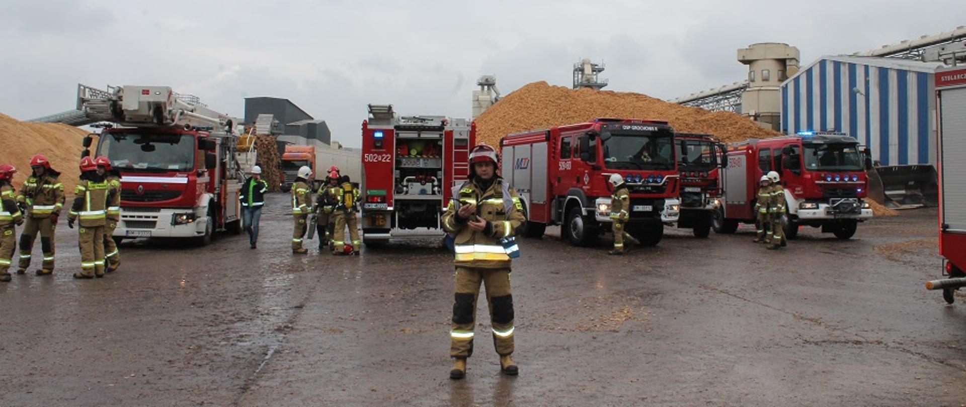 Fotografia przedstawia funkcjonariuszy przybyłych na ćwiczenia na terenie firmy Kronospan Mielec. Za strażakami widać cztery pojazdy ratowniczo-gaśnicze. W tle plac firmy i składowisko materiałów.
