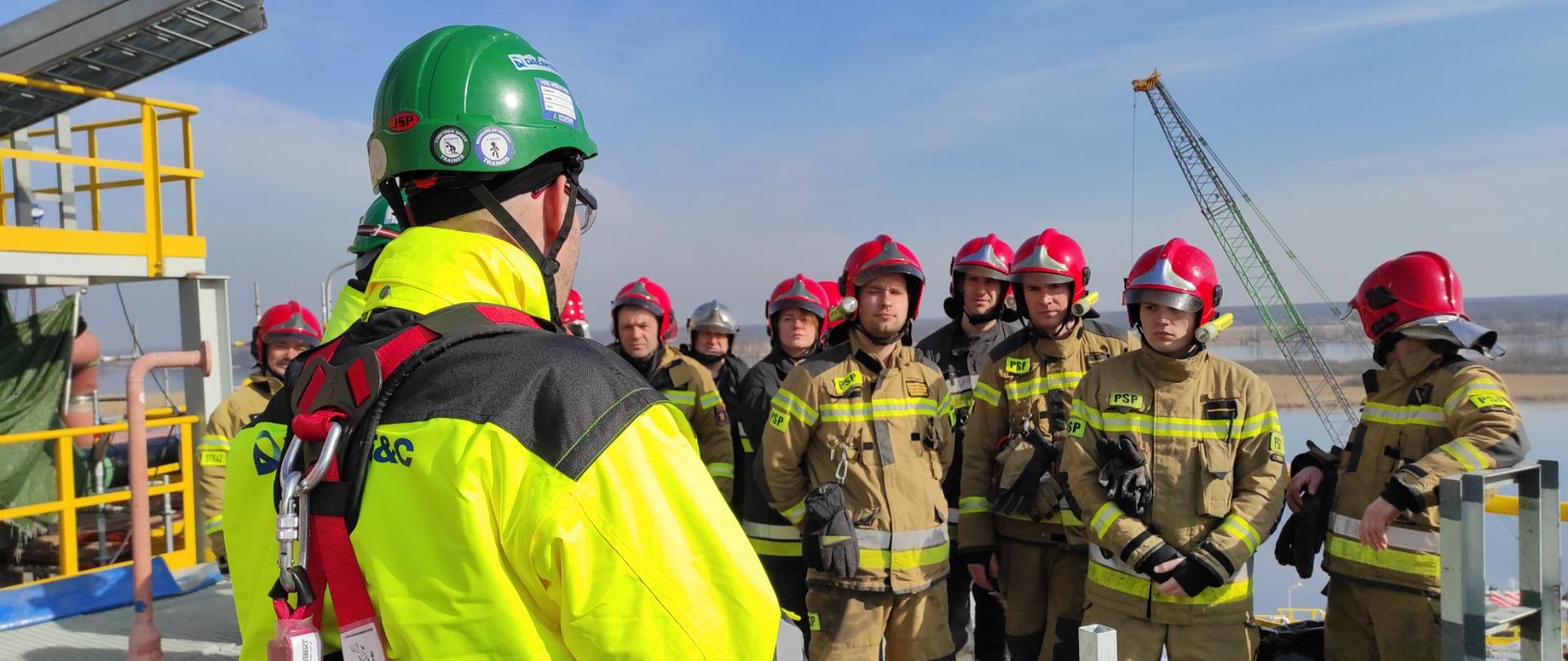 Na zdjęciu widoczna grupa stojących strażaków w piaskowych bojowych mundurach w czerwonych hełmach na głowie. opartych o żółta stalowa barierę na dachu zbiornika. W tle widoczne z dużej wysokości: odlegle tereny leśne i rozlewisko naturalnego zbiornika wodnego. Po lewej stronie zdjęcia widoczne plecy mężczyzny w jaskrawej jasnozielonej kurtce w zielonym kasku na głowie. Niebo błękitne z niewielkim zachmurzeniem.