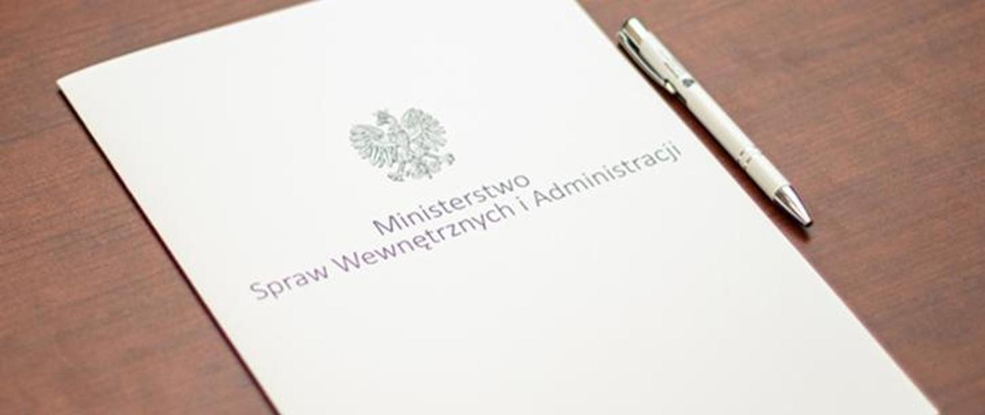 Biała teczka leży na biurku, z napisem Ministerstwo Spraw Wewnętrznych i Administracji i godłem. Z prawej strony leży biały długopis.