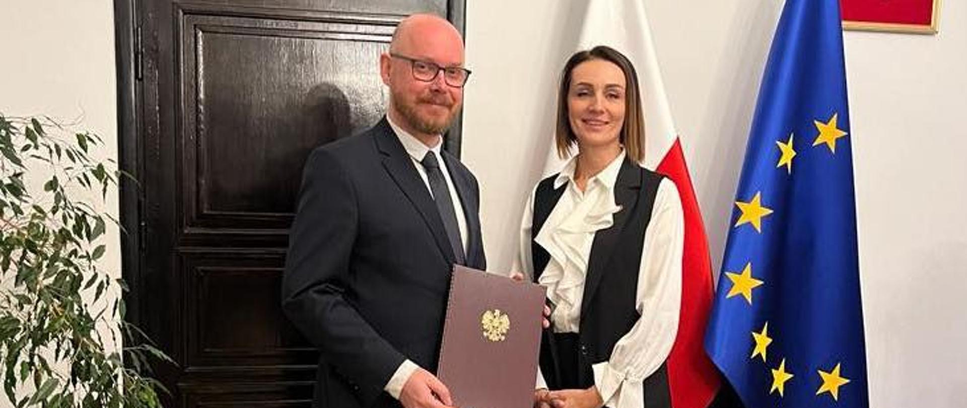 Gabinet wojewody pomorskiego, na tle flag polskiej i unijnej stoją dwie postaci, wojewoda wręcza teczkę z powołaniem 