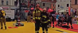 Na zdjęciu strażaków ubranych w ubrania bojowe, pozujących do zdjęcia na torze do konkurencji Firefit, w tle widać obsługę przygotowującą tor dla kolejnych startujących oraz widać wieżę po której wbiega się na początku konkurencji, wraz z kamienicami rynku Nowomiejskiego.