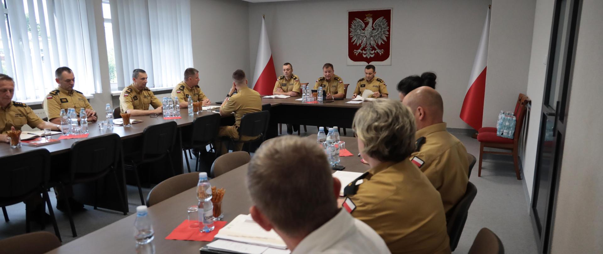 Na zdjęciu widać funkcjonariuszy PSP - Komendantów Wojewódzkich PSP, Komendantów Powiatowych i Miejskiego PSP oraz naczelników wydziałów KW PSP w Opolu w umundurowaniu służbowym zasiadających przy stole podczas narady kadry kierowniczej województwa opolskiego w sali Ośrodka Szkolenia KW PSP Opole. W tle godło i flaga Polski.
