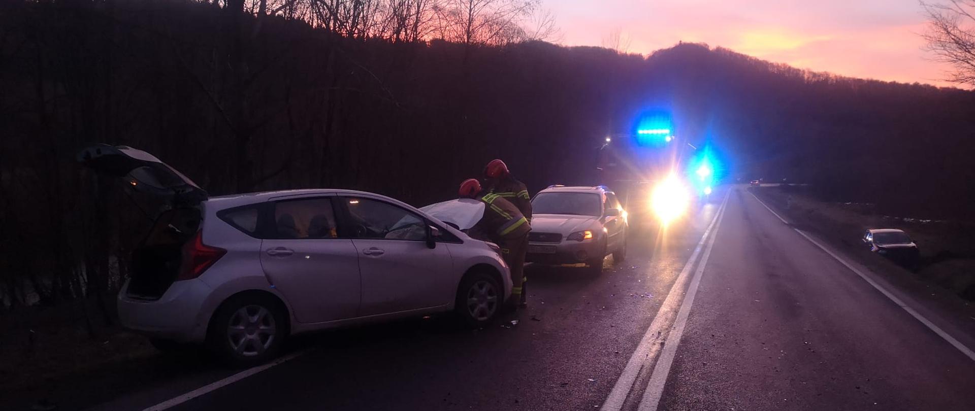 Zdjęcie przedstawia trzy samochody osobowe. Jeden ustawiony jest po prawej bokiem do drogi, drugi stoi przed nim a trzeci samochód znajduje się w rowie. Wzdłuż drogi ustawione są dwa samochody pożarnicze. Przy pierwszym pojeździe stoi dwóch strażaków. 