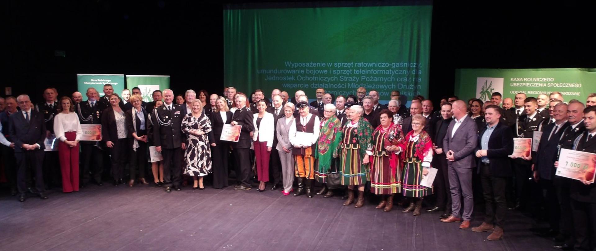 Grupa osób, kobiet i mężczyzn stojących na scenie na tle banerów KRUS. Część z nich w galowych mundurach strażackich trzyma w rękach vouchery z przyznaną kwotą dofinansowania.