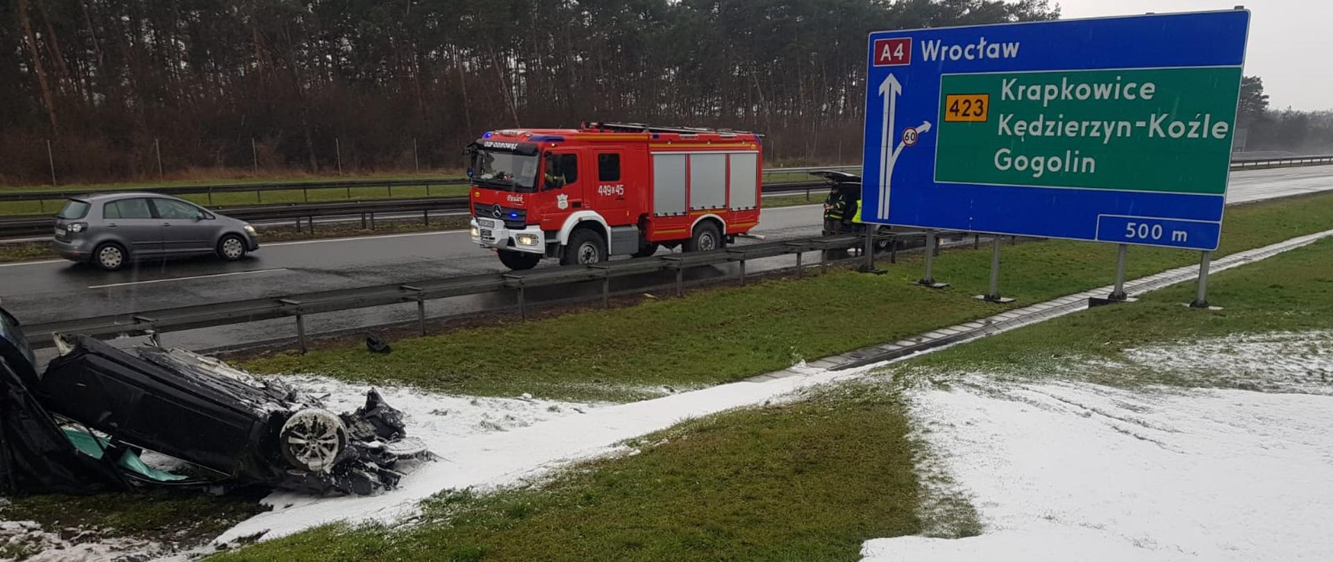 Zdjęcie przedstawia w lewym dolnym rogu rozbity samochód. W centralnej części widoczny samochód pożarniczy a po prawej stronie widoczny znak informacyjny. Na pierwszym planie trawa oraz położona na niej poduszka piany.