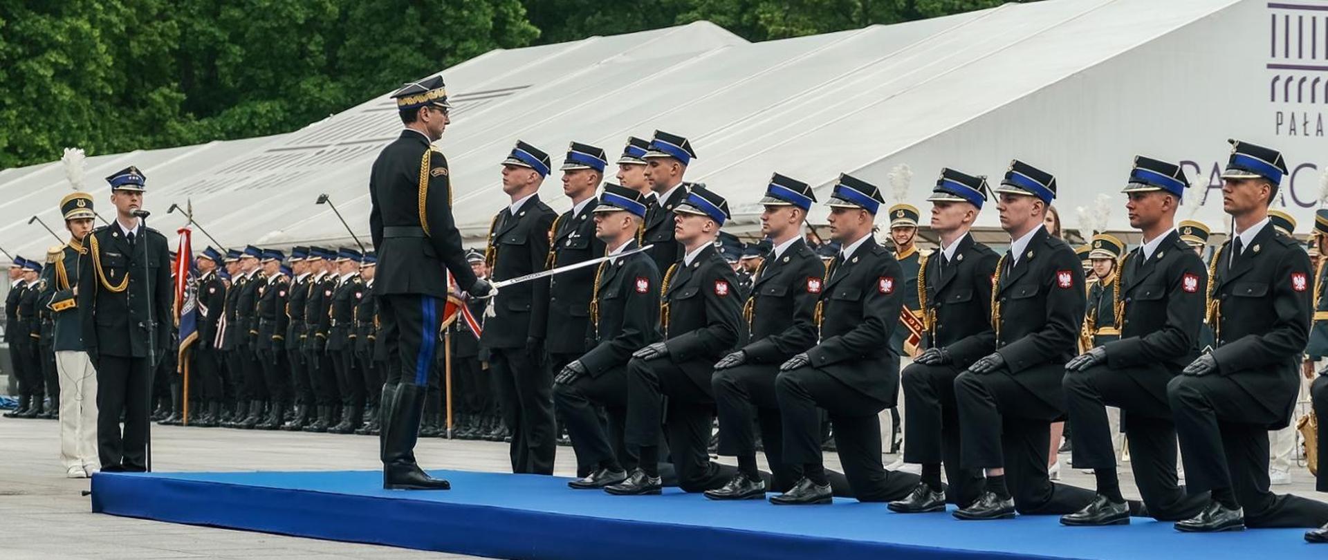 Zdjęcie przedstawia uroczystą ceremonię promocji na pierwszy stopień oficerski. Na niebieskim podest stoi dowódca, który szablą dotyka ramienia jednego z siedmiu klęczących przed nim funkcjonariuszy w czarnych mundurach galowych. W tle widoczne są rzędy umundurowanych osób, białe namioty oraz fragment napisu „PAŁAC”. Panuje podniosła, sformalizowana atmosfera.