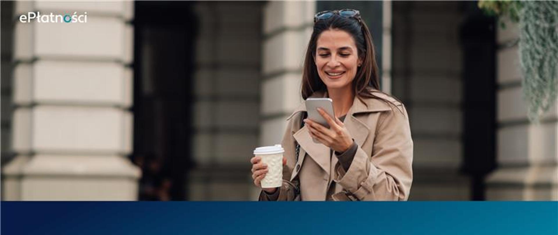 Uśmiechnięta kobieta z kawą w jednej ręce i telefonem w drugie, stoi przed budynkiem