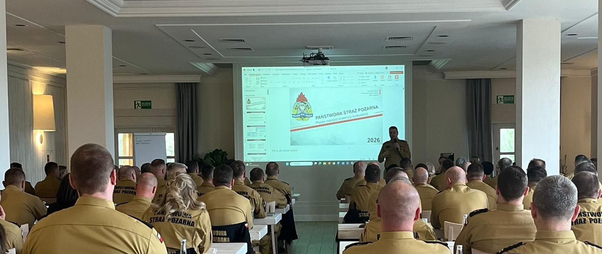 Sala konferencyjna wypełniona strażakami Państwowej Straży Pożarnej siedzącymi przy stołach i uczestniczącymi w naradzie. Na środku widoczny prelegent stojący przed ekranem z prezentacją PSP. 