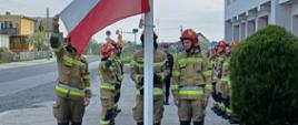 Zdjęcie przedstawia dwie zmiany służbowe stojące naprzeciw siebie podczas uroczystego podniesienia flagi na maszt, gdzie przy maszcie stoi trzech strażaków podnoszących flagę. Uroczystość ma miejsce na placu zewnętrznym Komendy Powiatowej Państwowej Straży Pożarnej w Krośnie Odrzańskim.