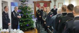Zdjęcie przedstawia spotkanie wigilijne w KP PSP Świdwin, które odbywa się na świetlicy. Strażacy są ubrani w mundury wyjściowe a ksiądz ich błogosławi.