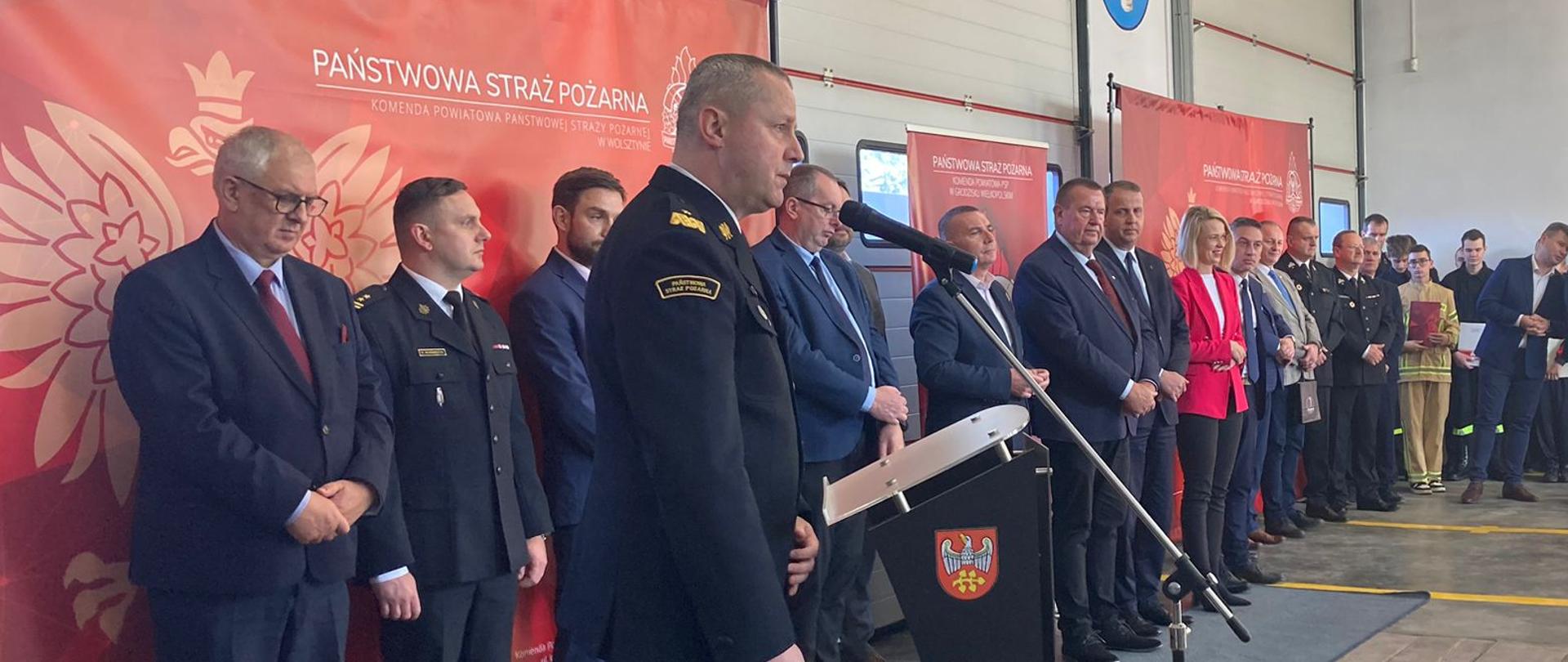 strażak generał stoi przy mównicy, za nim duża grupa osób składająca się ze strazaków i osób cywilnych