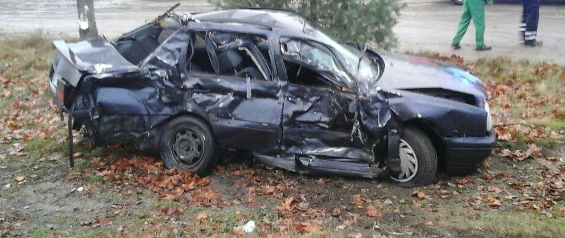 Samochód osobowy znajdując się w przydrożnym rowie. Całkowicie pognieciona karoseria z prawej strony pojazdu, wygięte koła, w oddali samochód strażacki i dwa cywilne pojazdy. 