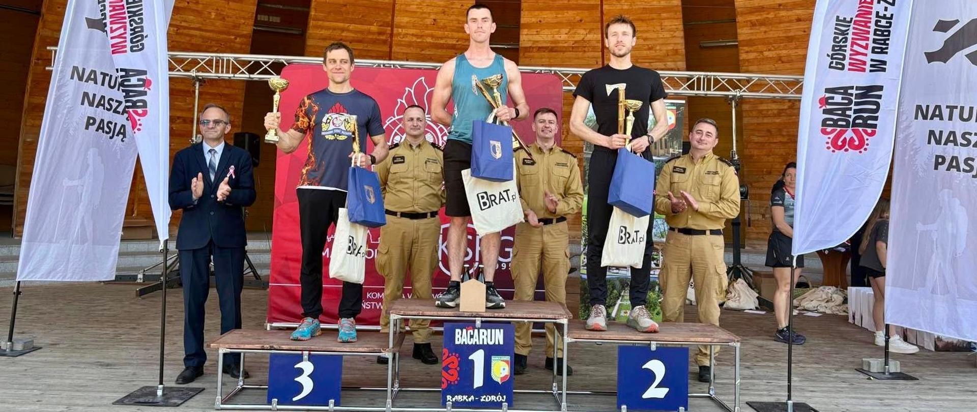 Zdjęcie przedstawia moment wręczania nagród. Na podium stoi trzech najlepszych zawodników. Zawodnicy ubrani są w stroje sportowe. W rękach trzymają puchary. Funkcjonariusze Państwowej Straży Pożarnej w ubraniach piaskowych składają gratulacje dla tych zawodników. Po lewej i prawej stronie podium widoczne są banery reklamowe. W tle widać drewnianą scenerię nawiązującą do miejsca w którym wręczane są nagrody. 
