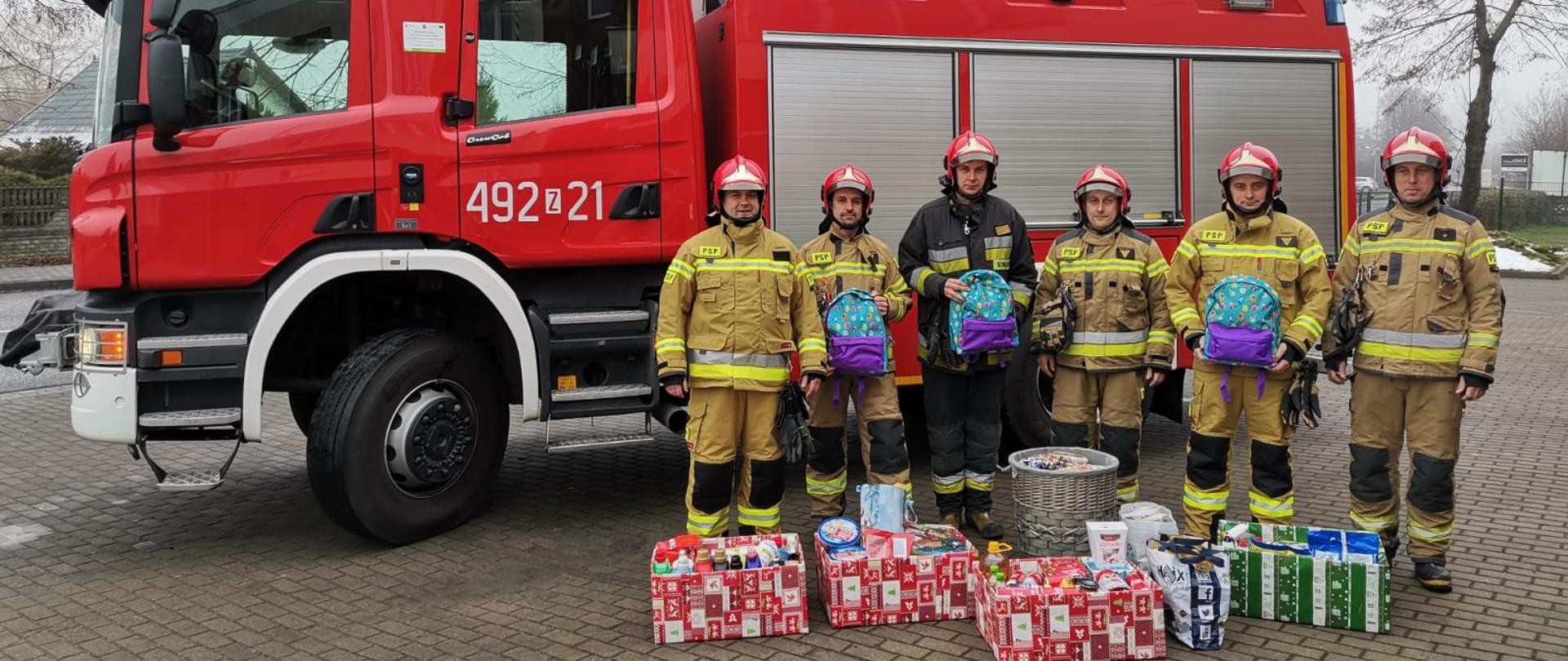 Na zdjęciu widzimy 6 strażaków stojących na tle samochodu pożarniczego. Strażacy są ubrani w umundurowanie specjalne i hełmy. Naprzeciwko strażaków ustawione na ziemi są świąteczne paczki i prezenty.