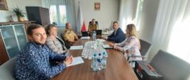 Sześć osób siedzi wokół konferencyjnego stołu w urzędowym biurze. Dwóch uczestników po lewej stronie ma beżowe mundury strażackie z naszywkami i dystynkcjami. Pozostałe osoby są w eleganckiej odzieży biurowej. Na stole leżą dokumenty, butelki wody i biały koronkowy bieżnik. Przed osobą siedzącą na końcu stołu stoi otwarty laptop. W tle widoczna jest polska flaga, godło Polski na ścianie, szafka z segregatorami oraz roślina doniczkowa. Scena przedstawia formalne spotkanie robocze związane z interdyscyplinarnym projektem dotyczącym ochrony zdrowia strażaków i monitorowania zagrożeń środowiskowych.