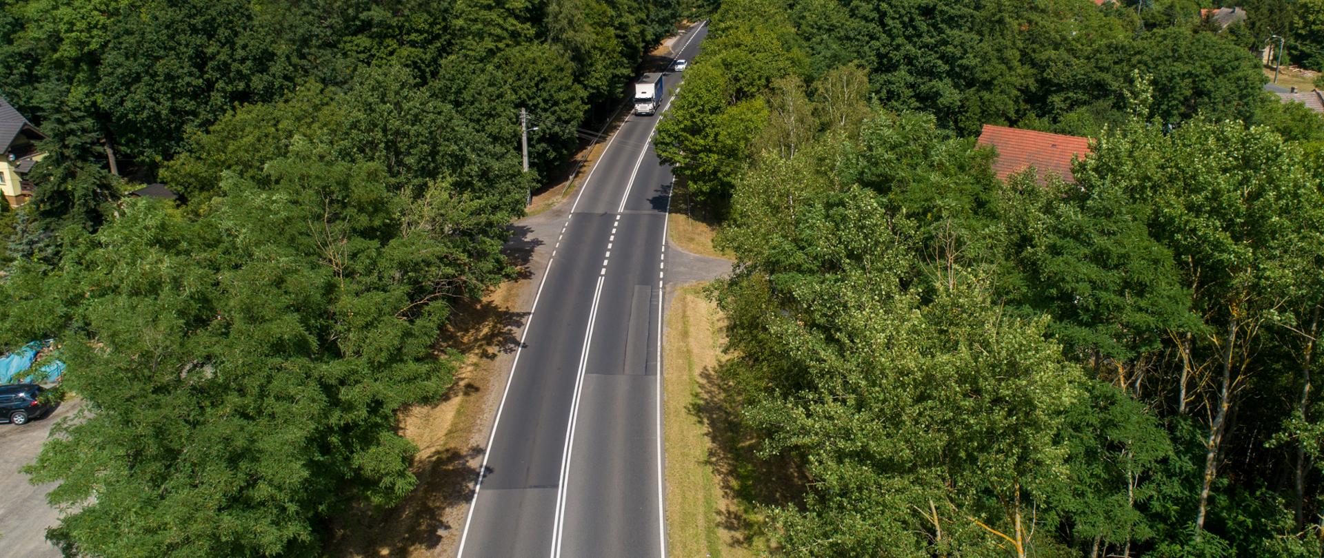 Droga jednojezdniowa z lotu ptaka, w otoczeniu lasów i pojedynczych zabudowań jednorodzinnych.