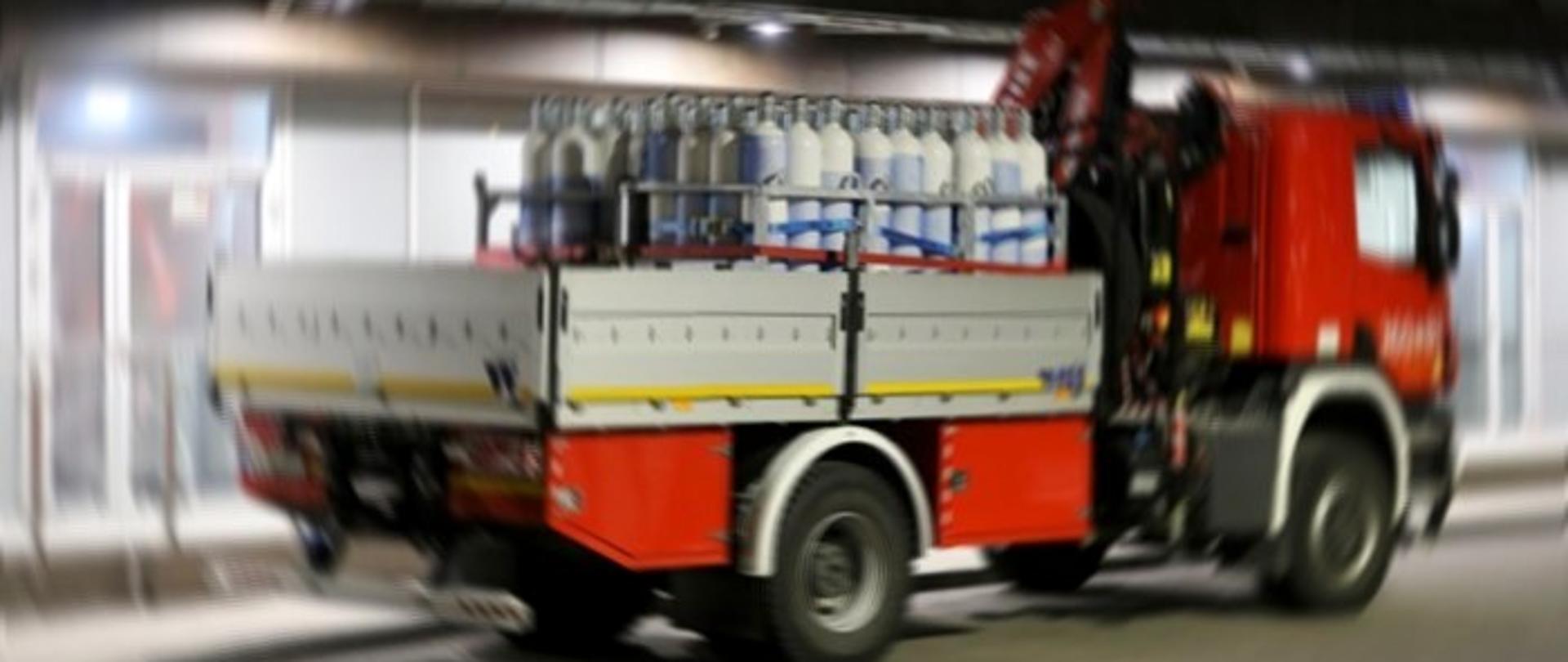 Na zdjęciu widoczny jest pożarniczy samochód specjalny kwatermistrzowski stojący na przed budynkiem szpitalnym. W przestrzeni załadunkowej pojazdu znajduje się bateria butli z tlenem, umieszczonych w specjalnej konstrukcji przeznaczonej do ich transportu.