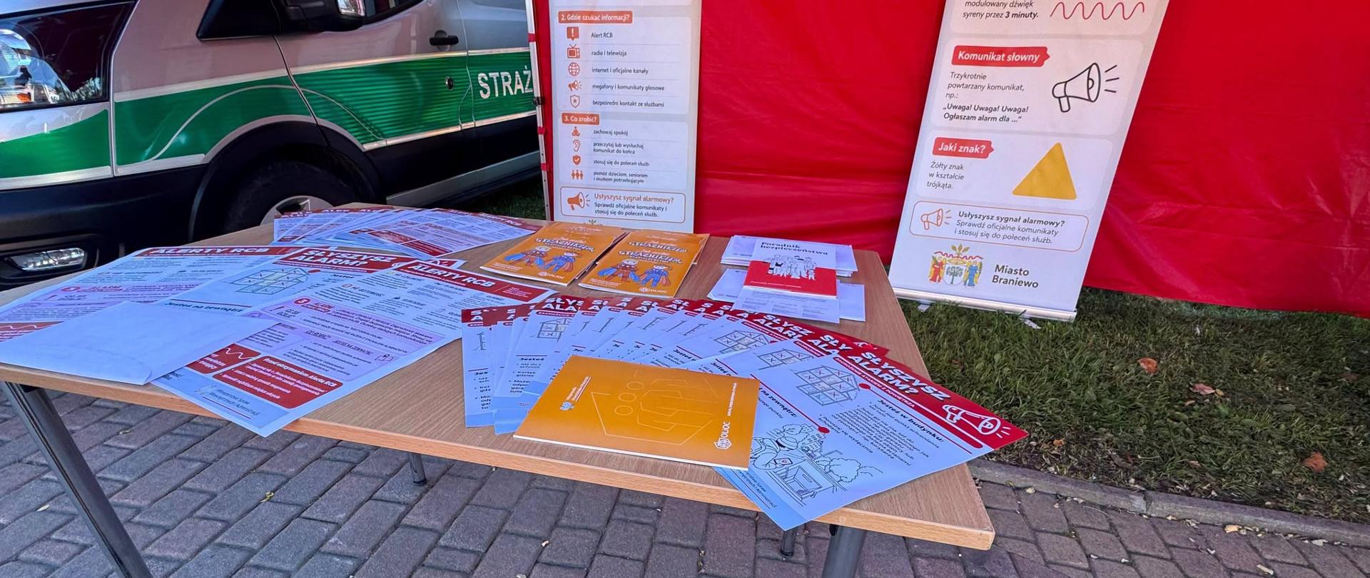 Na fotografii widoczne jest stoisko informacyjne poświęcone zasadom alarmowania i właściwego reagowania w sytuacjach zagrożenia. Na stole przygotowano materiały edukacyjne, ulotki i broszury dotyczące m.in. sygnałów alarmowych, komunikatów ostrzegawczych oraz zasad postępowania w przypadku wystąpienia zagrożenia. Obok ustawiono plansze informacyjne przedstawiające sposób ogłaszania alarmu i źródła, z których można uzyskać wiarygodne informacje. W tle widoczny jest pojazd Straży Granicznej.