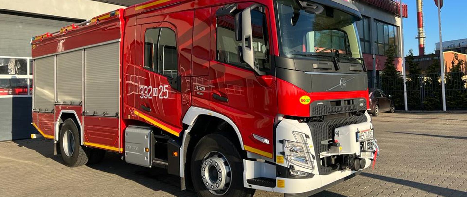 Zdjęcie przedstawia ciężki pojazd ratowniczo - gaśniczy GCBA 5/32. Pojazd w kolorze czerwonym wraz ze srebrnymi i białymi elementami zabudowy. Samochód ustawiony przed garażami Jednostki Ratowniczo – Gaśniczej nr 2 w Radomiu.