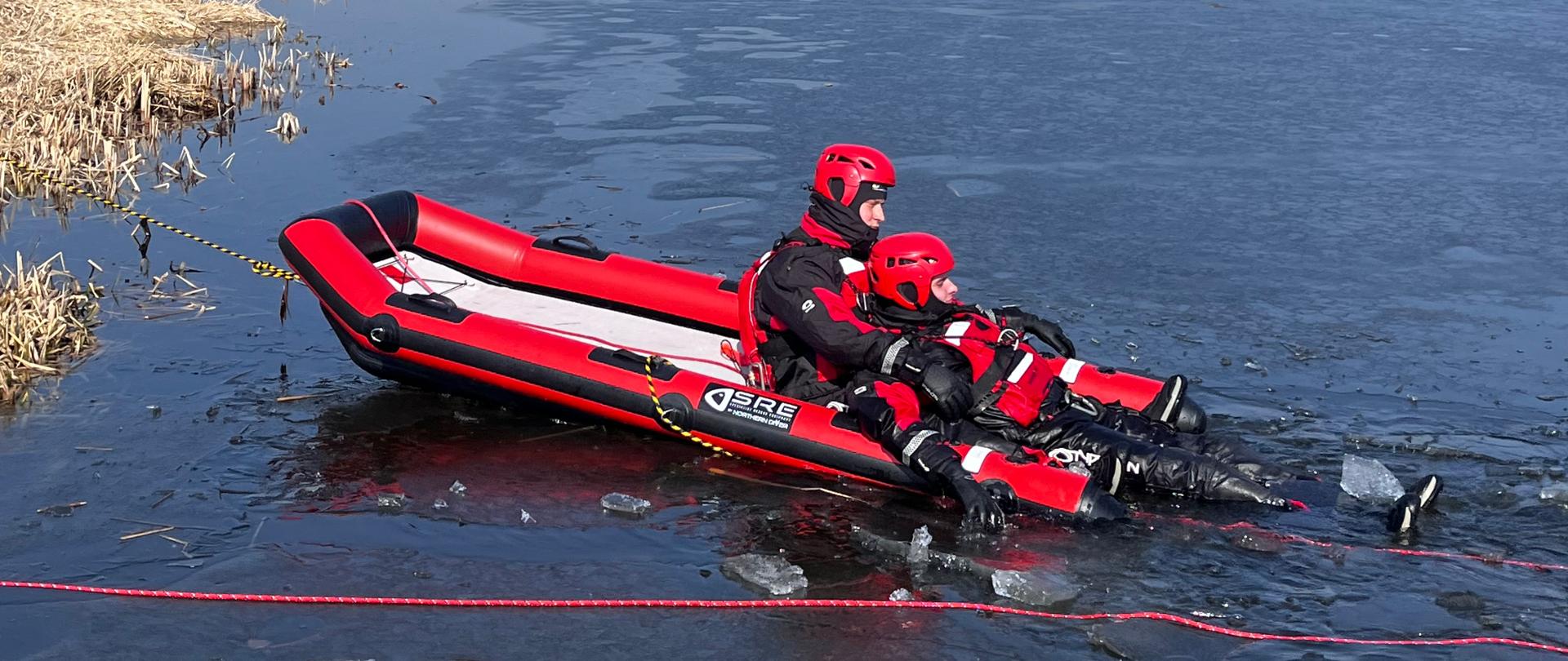 Dwóch ratowników w czerwonych kombinezonach i kaskach siedzi w pontonie na zamarzniętym jeziorze podczas ćwiczeń z ratownictwa lodowego. Wokół widać pękający lód, linę asekuracyjną oraz brzeg z trzciną i drzewami.