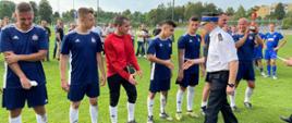 Zwycięska drużyna w niebieskich strojach stoi w rzędzie, a komendant wojewódzki gratuluje piłkarzom ściskając im dłonie.
