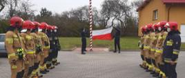 Strażacy stoją w dwóch rzędach na przeciwko siebie, uroczyście podnosząc flagę Rzeczypospolitej Polskiej na maszt.
