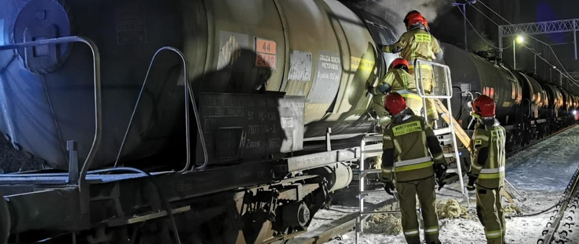 Cysterny kolejowe. Przy jednej z nich, gdzie wydobywa się dym, znajduje się podest roboczy na którym pracuje trzech strażaków. Dwóch strażaków stoi przy podeście. Na podłożu jest śnieg i reszki izolacji cysterny oraz linia gaśnicza szybkiego natarcia.