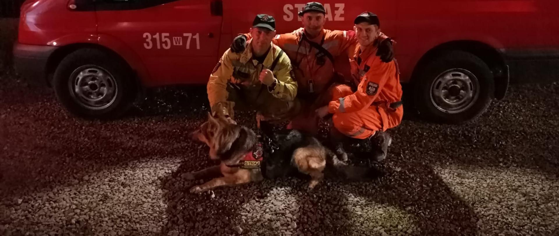 Na zdjęciu widać trzech członków specjalistycznej grupy poszukiwawczo ratowniczej wraz z psem ratowniczym Gines. Zdjęcie wykonane w porze nocnej. Ratownicy znajdują się przed pojazdem, którym transportuje się psy ratownicze.