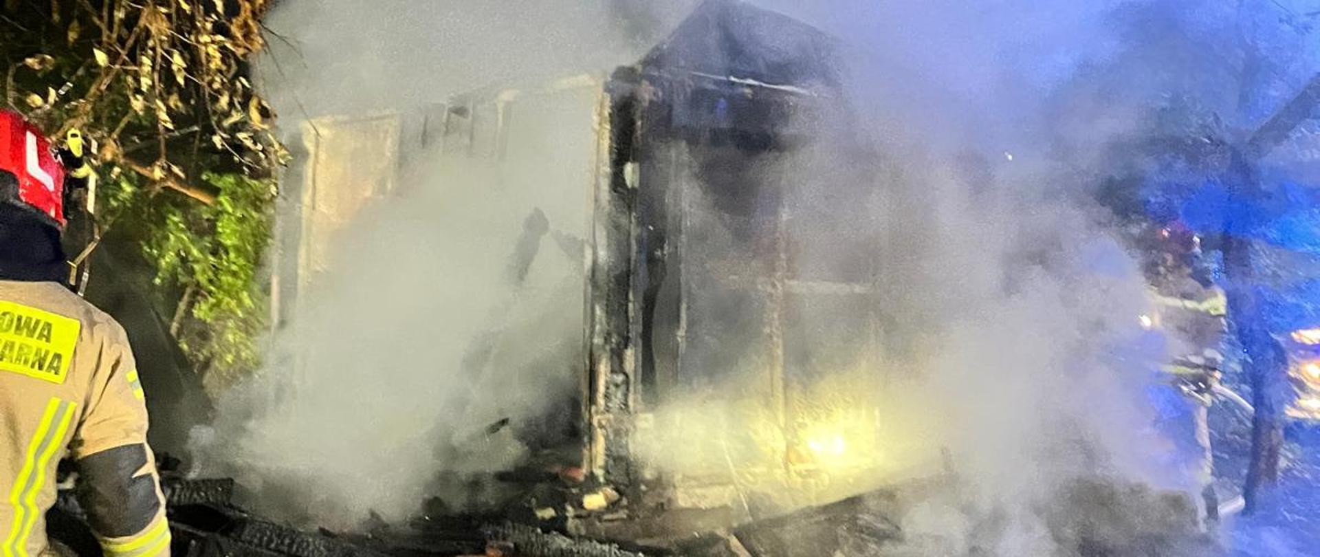 Zdjęcie przedstawia szczątki spalonej altanki działkowej a z lewej strony widać strażaka.