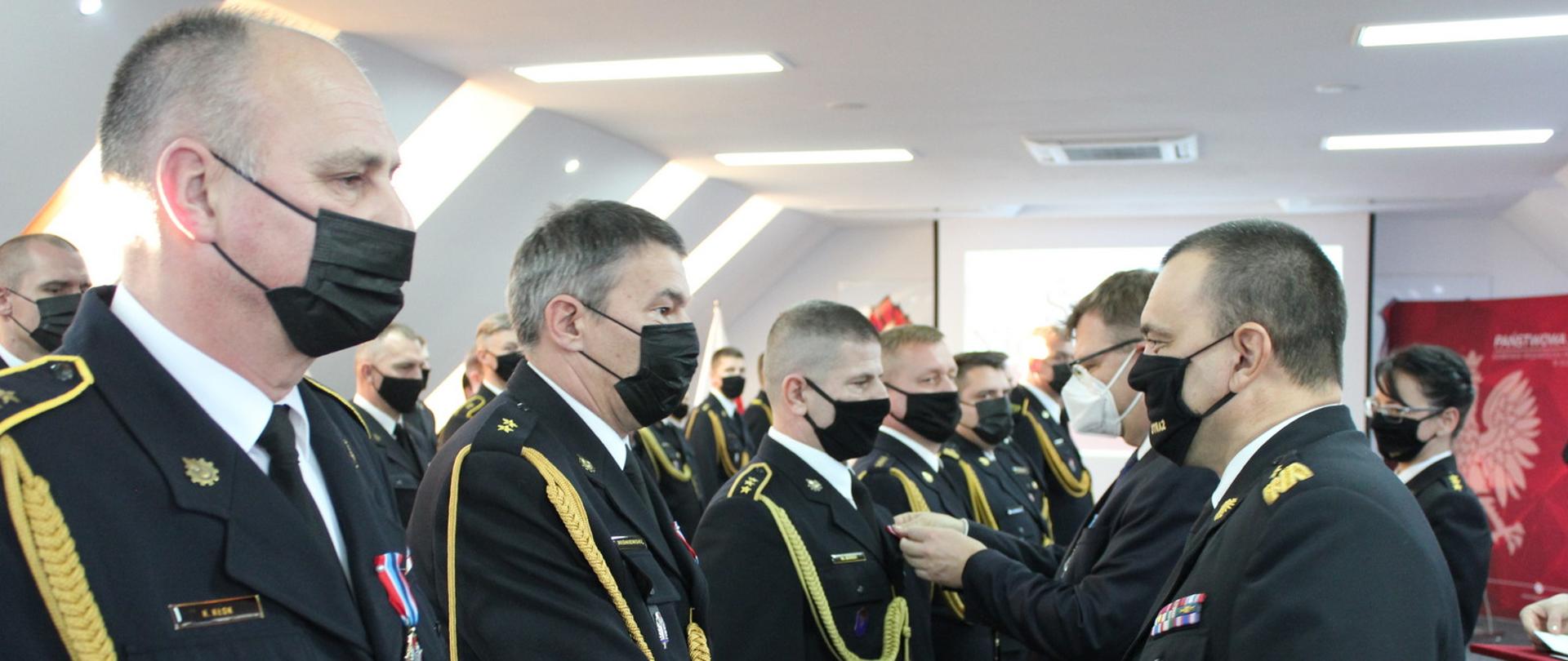Zdjęcie zrobione w Sali konferencyjnej Ośrodka Szkolenia PSP w Gutkowie. Funkcjonariusze ubrani w czarny mundur galowy ze złotym sznurem. Komendant wojewódzki podaje rękę wyróżnionemu funkcjonariuszowi.
