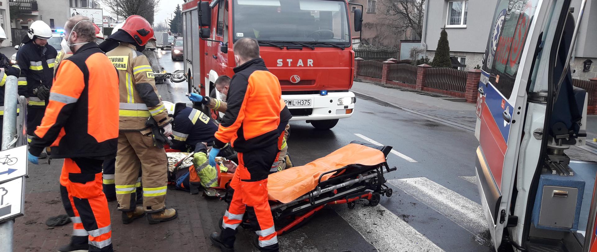 Po prawej stronie karetka. W środku strażacy oraz ratownicy medyczni zajmują się osobą poszkodowaną.