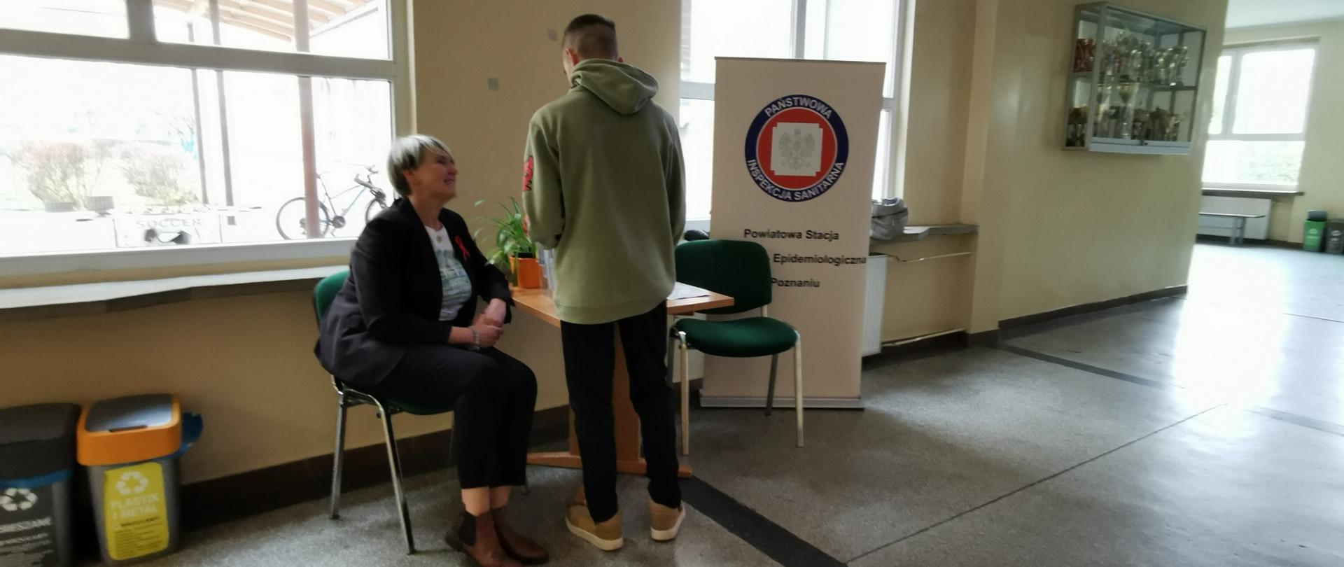 Na korytarzu szkolnym uczeń stoi przy stoliku, obok siedzi pracownica Powiatowej Stacji Sanitarno-Epidemiologicznej w Poznaniu. W tle baner z logo Inspekcji Sanitarnej.