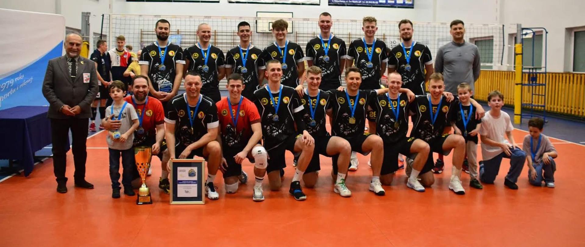 Drużyna siatkarska Centralnej Szkoły PSP pozująca do wspólnego zdjęcia na hali sportowej. Zawodnicy ubrani są w czarne stroje sportowe i mają na szyjach medale. Część z nich stoi, a część klęczy w pierwszym rzędzie. Na środku, przed zawodnikami, ustawiony jest puchar oraz dyplom potwierdzający zdobycie tytułu. Obok drużyny stoją także inne osoby, w tym mężczyzna w garniturze oraz kilkoro dzieci. W tle widoczna jest siatka do siatkówki oraz ściany hali sportowej z banerami reklamowymi. Całość przedstawia moment po zakończeniu rozgrywek i celebrację zwycięstwa drużyny