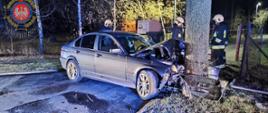 Zdjęcie przedstawia zniszczony samochód po uderzeniu w drzewo oraz strażaków
